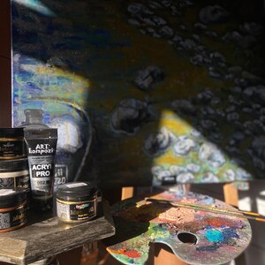 May include: A close-up of a set of acrylic paint bottles and jars, including a black bottle labeled "Art Compozi Acrylic Pro 002". The bottles are on a wooden table next to a paint palette with various colors of paint.