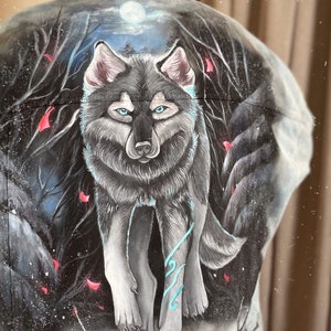 May include: A denim jacket featuring a hand-painted design of a wolf in a dark forest setting. The wolf is grey and white with blue accents. The background is dark with red and white accents.