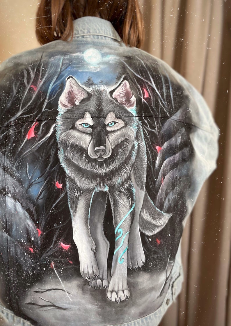 May include: A denim jacket featuring a hand-painted design of a wolf in a dark forest setting. The wolf is grey and white with blue accents. The background is dark with red and white accents.