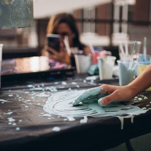 May include: A close-up of a hand covered in blue paint, creating a swirl pattern on a black surface. There are several cups and containers of paint in the background.