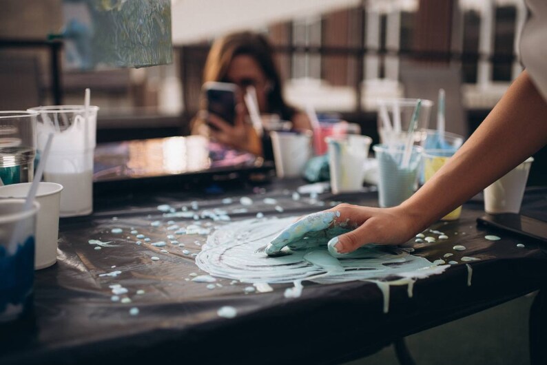 May include: A close-up of a hand covered in blue paint, creating a swirl pattern on a black surface. There are several cups and containers of paint in the background.