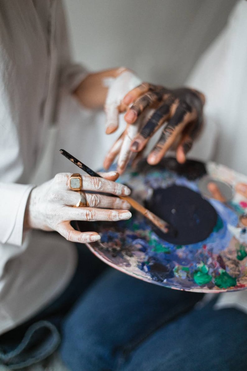 May include: A person's hands holding a paint palette with a variety of colors, including blue, green, and yellow. A paintbrush is resting on the palette.