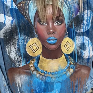 Op de afbeelding: Een spijkerjack met een geschilderd ontwerp van een vrouw met blauwe en gouden sieraden, een blauwe hoofddoek en blauwe en witte verfspatten.