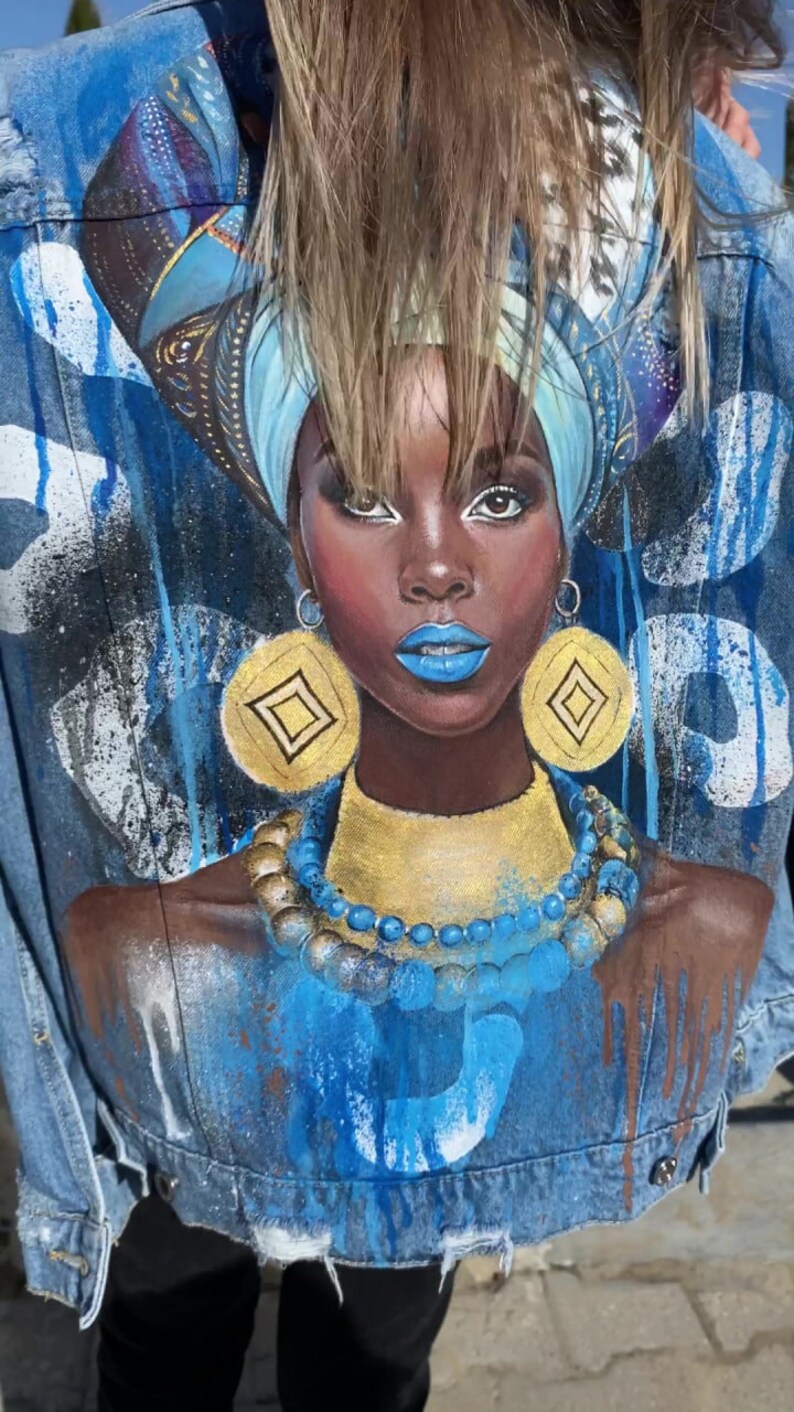 Op de afbeelding: Een spijkerjack met een geschilderd ontwerp van een vrouw met blauwe en gouden sieraden, een blauwe hoofddoek en blauwe en witte verfspatten.