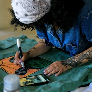 May include: A person is drawing on a teal fabric with black and white markers. The fabric has a colorful design of two faces with orange, yellow, green, and black colors. The person has a dragon tattoo on their arm.