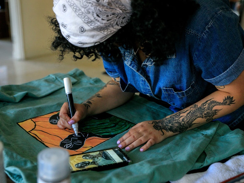 May include: A person is drawing on a teal fabric with black and white markers. The fabric has a colorful design of two faces with orange, yellow, green, and black colors. The person has a dragon tattoo on their arm.