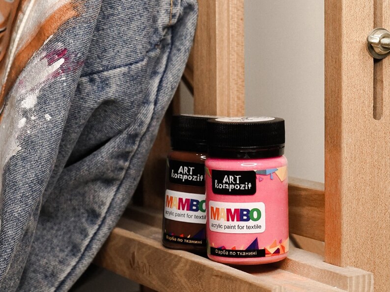 May include: Two bottles of acrylic paint for textile, labelled "MAMBO" in a colourful font. The bottles are brown and pink and have black lids. The text on the labels reads "ART kompozit" and "acrylic paint for textile" in Russian.