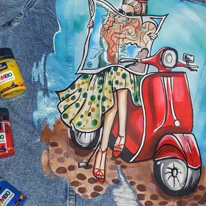 May include: A denim jacket featuring a painted design of a woman riding a red scooter. The woman is holding a map and wearing a polka dot dress. The jacket has three paint bottles on it, labelled "Art Lapatch Mambo acrylic paint for fabric".