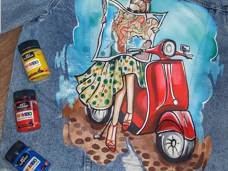 May include: A denim jacket featuring a painted design of a woman riding a red scooter. The woman is holding a map and wearing a polka dot dress. The jacket has three paint bottles on it, labelled "Art Lapatch Mambo acrylic paint for fabric".