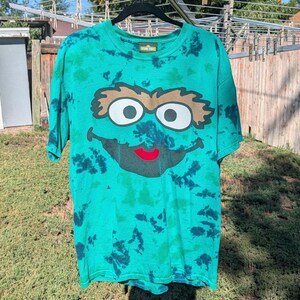 Könnte beinhalten: Ein kurzärmeliges T-Shirt mit Rundhalsausschnitt und einem Batikmuster in leuchtendem Grün und dunklem Petrol. Auf der Vorderseite befindet sich ein großes Motiv des Sesame Street Charakters Oscar aus der Mülltonne mit rotem Mund und Etikett.