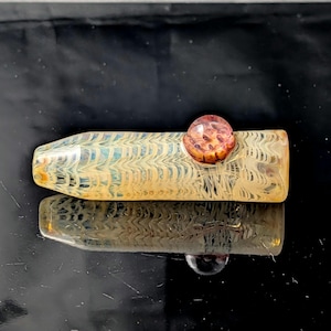 May include: A glass pipe with a yellow and white patterned design. The pipe has a rounded, amber-colored sphere on top. The pipe is reflected on a black surface.