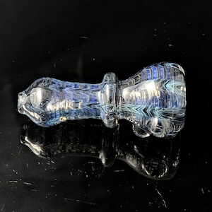 Pode incluir: Um cachimbo de vidro transparente com redemoinhos azuis e verde-azulados. O cachimbo tem uma tigela bulbosa e um caule cilíndrico. O design apresenta uma série de linhas e curvas paralelas, criando uma aparência texturizada. O cachimbo está sobre uma superfície preta reflexiva.