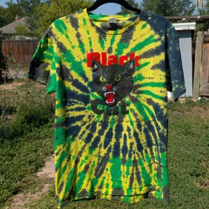May include: A short-sleeve crew neck t-shirt featuring a yellow, green, and black tie-dye pattern. The center graphic displays a black cat head with green eyes and a red mouth, with the red text Black Cat and the phrase Black Cat the Best you can get.