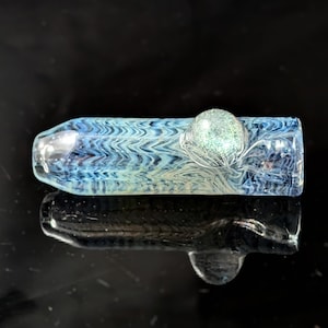 Pode incluir: Um cachimbo de vidro transparente com um padrão azul e branco em espiral. Uma conta redonda, brilhante, verde claro é colocada no meio do cachimbo. O cachimbo é refletido na superfície preta abaixo.