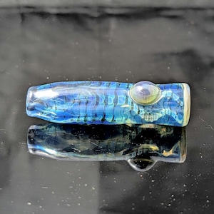 May include: A cylindrical glass pipe featuring a deep blue and teal color palette with a repeating wave-like pattern throughout the body. The piece includes a small, raised circular glass bead on the side and a clear, flat mouthpiece end.