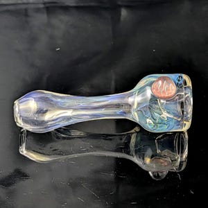 Könnte beinhalten: Eine klare Glaspfeife mit Kopf und Stiel. Die Pfeife hat blaue und grüne Akzente, mit einer rötlich-braunen Kugel auf dem Kopf. Das Glas ist reflektierend, und die Pfeife liegt auf einer dunklen Oberfläche.