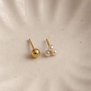 May include: Two gold stud earrings displayed on a white, scalloped-edge dish with a gold rim. One earring is a simple gold ball stud, and the other features three small, white pearls. The dish is set against a neutral background.