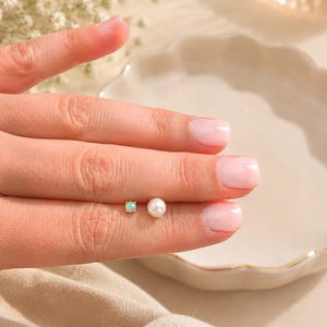 May include: A hand displays two earrings: a small square stud with a light green stone and a round pearl stud. The hand is positioned near a white, scalloped-edge dish. Soft lighting and a neutral background enhance the jewelry's delicate appearance.