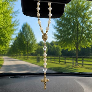 Op de afbeelding: Een rozenkrans hangend aan de achteruitkijkspiegel van een auto met witte kralen, heldere gefacetteerde kristallen kralen en goudkleurige metalen accenten. De rozenkrans bevat een centrale ovale medaille met een figuur en een gouden kruisbeeld.