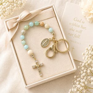 Puede incluir: Un llavero de rosario con cuentas de color azul claro, beige y blanco, detalles dorados y un colgante de cruz. El llavero incluye un cierre dorado y un pequeño dije ovalado. El texto "God is within her, she will not fall. PSALM 46:5" es visible en una tarjeta.