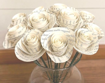 Puede incluir: Un ramo de rosas de papel hechas con páginas de libros, exhibido en un jarrón de vidrio transparente. Las rosas de color crema tienen texto visible y los tallos son verdes. Un artículo decorativo único y artesanal.