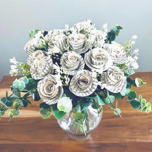 May include: A bouquet of paper roses made from book pages, arranged in a clear glass vase. The roses are white with visible text, complemented by green foliage and small white flowers. The arrangement sits on a wooden surface.