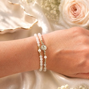 May include: A set of two stretch bracelets featuring small, smooth, pale pinkish-white beads interspersed with polished gold-tone metallic beads. One bracelet includes a circular gold-tone charm embossed with a detailed religious figure.