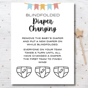 May include: A white card with black text and illustrations. The text reads "BLINDFOLDED Diaper Changing". The text continues, "REMOVE THE BABY'S DIAPER AND PUT A NEW DIAPER ON WHILE BLINDFOLDED. EVERYONE ON YOUR TEAM TAKES A TURN UNTIL ALL HAVE CHANGED A DIAPER. THE FIRST TEAM TO FINISH WINS!" Three black and white illustrations of diapers with safety pins are at the bottom of the card.