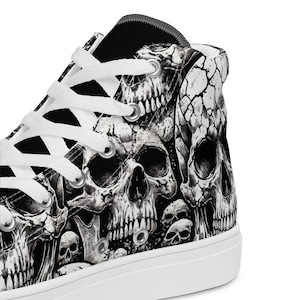 Op de afbeelding: Hoge canvas sneakers met een zwart-wit schedelpatroon. Het ontwerp bevat meerdere schedels van verschillende groottes, wat een gotische esthetiek creëert. De schoenen hebben witte veters en een witte rubberen zool.
