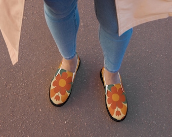 Pode incluir: Um par de sapatos slip-on com um padrão floral. Os sapatos apresentam um fundo creme com grandes flores laranja e amarelas, folhas verdes e detalhes vermelhos. Solas pretas, usados com jeans azuis. Estilo retro.