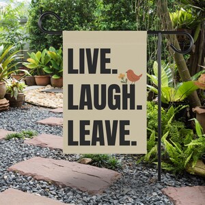 May include: A beige garden flag with the words "LIVE. LAUGH. LEAVE." in black block letters. A small orange bird and yellow flowers are to the right of the word "LEAVE." The flag is hanging on a black metal garden stake.