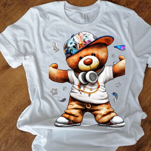 Pu&ograve; includere: Maglietta bianca con una grafica di un orsacchiotto marrone che indossa un cappello colorato, cuffie e pantaloni marroni. L'orso sta flesso i muscoli.