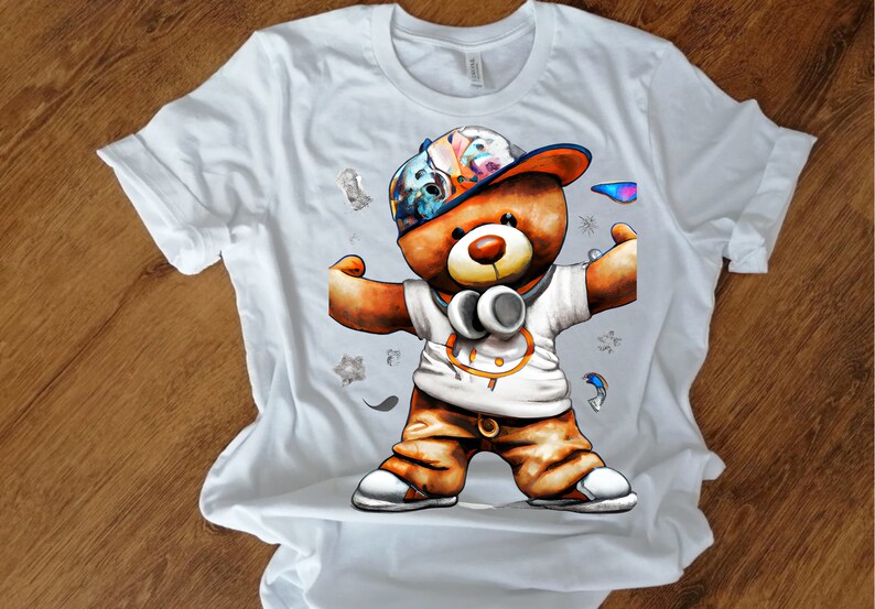 Pu&ograve; includere: Maglietta bianca con una grafica di un orsacchiotto marrone che indossa un cappello colorato, cuffie e pantaloni marroni. L'orso sta flesso i muscoli.