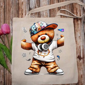 Pu&ograve; includere: Una borsa tote beige con un'illustrazione colorata di un orsacchiotto che indossa un cappello, cuffie e una camicia bianca con una cintura marrone. L'orso &egrave; in piedi con le braccia distese.