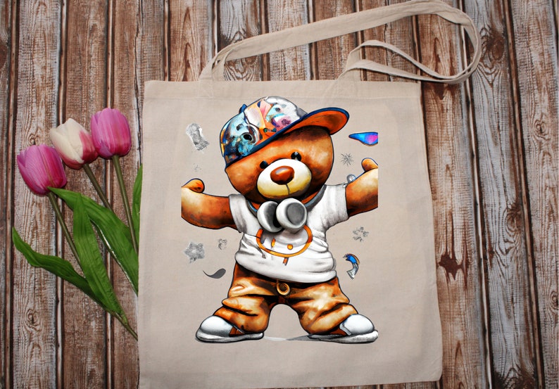 Pu&ograve; includere: Una borsa tote beige con un'illustrazione colorata di un orsacchiotto che indossa un cappello, cuffie e una camicia bianca con una cintura marrone. L'orso &egrave; in piedi con le braccia distese.