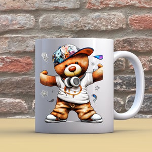 Pu&ograve; includere: Tazza in ceramica bianca con un'illustrazione colorata di un orso che indossa un cappello e delle cuffie. L'orso &egrave; in piedi con le braccia distese e indossa una camicia bianca con un cerchio nero. La tazza ha un manico bianco.