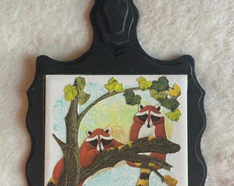 Può includere: Una placca da parete in legno nero con bordo smerlato e un foro per appenderla. Il centro presenta una piastrella quadrata con un'illustrazione in stile collage di due procioni rossi e neri appollaiati su un ramo con foglie verdi.