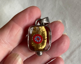 Può includere: Un ciondolo rettangolare in vetro incastonato in una cornice di metallo color argento. Il vetro presenta una perla millefiori centrale con motivi rossi, bianchi e blu, circondata da accenti in foglia dorata testurizzata in un corpo in vetro ambrato.