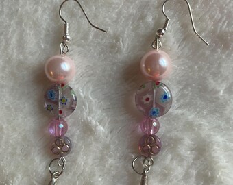 Può includere: Un paio di orecchini pendenti con ganci color argento, ciascuno caratterizzato da una grande perla rosa pallido, una perlina di vetro trasparente con piccoli motivi floreali blu e rossi, una perlina rosa traslucida e un ciondolo argentato a goccia.