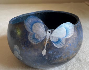Può includere: Una ciotola arrotondata con una finitura esterna maculata blu scuro e nera. La superficie presenta una farfalla dipinta a mano in azzurro e bianco con delicati dettagli sulle ali. Una farfalla più piccola è dipinta sul lato inferiore sinistro.