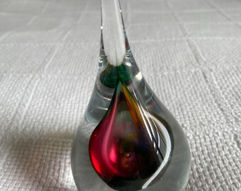 Può includere: Un fermacarte in vetro trasparente a forma di goccia. L'interno presenta un vivace mix di colori, tra cui rosso, giallo e verde. Il fermacarte ha una superficie liscia e lucida e una parte superiore lunga e appuntita.