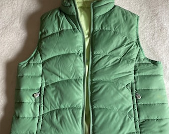 Può includere: Un gilet imbottito reversibile con esterno verde salvia con cuciture orizzontali e interno verde lime chiaro. Il gilet presenta una chiusura frontale con zip, un colletto rialzato e due tasche laterali con tiretti. Il logo LLBean è visibile.