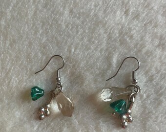 Può includere: Un paio di orecchini a gancio color argento con ciondoli pendenti. Ogni orecchino include una piccola perla verde a forma di fiore, una goccia trasparente sfaccettata simile al cristallo e un gruppo di piccole sfere metalliche color argento.
