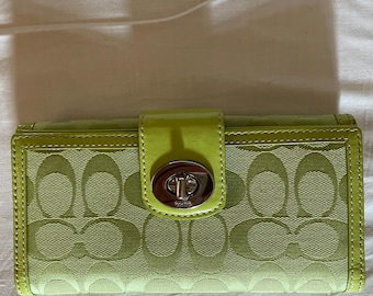 Può includere: Un portafoglio rettangolare verde lime con un motivo a logo circolare ripetuto su un corpo in tessuto testurizzato. Il portafoglio include una chiusura con cinturino in pelle verde lime liscia fissata da un fermaglio girevole in metallo argentato.