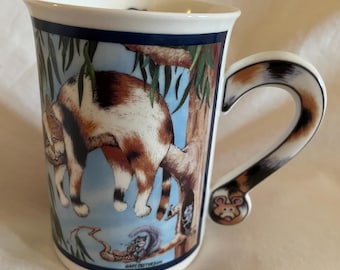 Può includere: Una tazza in ceramica bianca con un'illustrazione colorata di un gatto appeso a un ramo d'albero. Il manico della tazza è a forma di coda di gatto e ha una piccola testa di topo all'estremità. La tazza è decorata con un bordo blu e la firma dell'artista, "Gary Patterson".