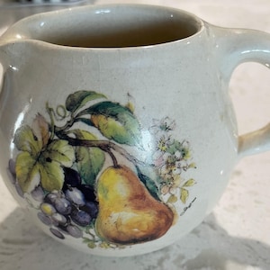 May include: A white ceramic pitcher with a handle, featuring a painted design of a pear, grapes, and leaves. The pitcher has a slightly rounded shape and a smooth, glossy finish.