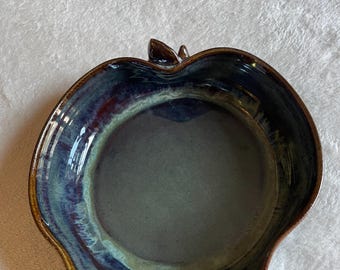 Può includere: Una ciotola in ceramica a forma di mela con una smaltatura lucida che presenta sfumature miste di blu profondo, viola e verde. L'interno ha un centro liscio grigio scuro, mentre il bordo e il piccolo dettaglio del gambo sono marroni.