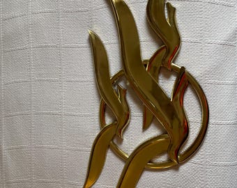 Può includere: Spilla decorativa in metallo color oro con un design astratto. La spilla presenta forme curve a forma di fiamma e un elemento circolare, il tutto con una finitura lucida. Il design è posto su uno sfondo bianco.