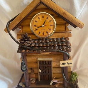 May include: A wooden cabin-shaped clock with a quartz clock face and a sign that reads "Cabin Fever". The clock is decorated with pine cones, a small door, and a lantern.