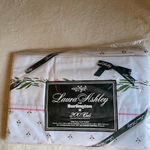 May include: A Laura Ashley for Burlington full flat sheet in white with a pink and green floral pattern. The packaging features a black label with white text, 200 thread count, and dimensions of 81 x 96 inches, made of 50% cotton and 50% polyester.